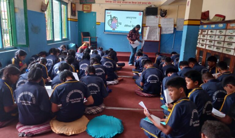 The participants are in the school hall sitting on cushions on the floor. They are facing towards the screen with their back at the camers. The participants are filling the pre-test questionnaire. The screen reads GBV and disability.